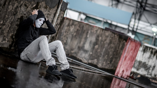 Mystery hoodie man in white mask feeling stressed sitting in the rain on rooftop of abandoned building. Bipolar disorder or Major depressive disorder. Depression concept