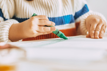 Fototapeta premium Cropped image of cute little girl paints with green oil pencil sitting at white desk at home. Image of pretty preschool kid draws on paper in the kindergarten. People, childhood and education concept