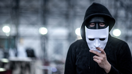 Mystery hoodie man with broken black mask holding white mask in his hand. Anonymous social masking...