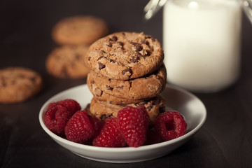 Chocolate chip cookies on white plate dark old wooden table with red raspberry and milk. Delicious breakfast.