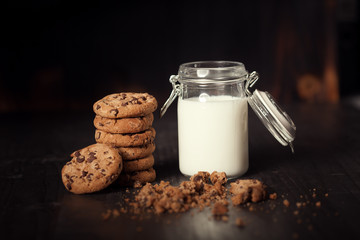 A stack of chocolate chip cookies with milk on dark old wooden table. Delicious snack.