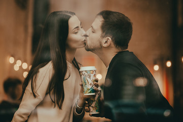 Happy lovers guy and girl are kissing on the windowsill in a cafe and holding cups in their hands