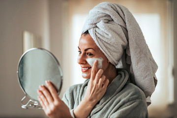 Beautiful smiling woman holding mirror and applying face cream at home. - obrazy, fototapety, plakaty