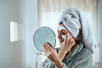 Smiling young woman holding a mirror and applying face cream. - obrazy, fototapety, plakaty