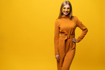 Excited young woman in orange dress, widely smiling, looking at camera. Isolated on yellow background