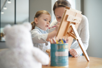 Mother teaching young daughter to draw on easel at home