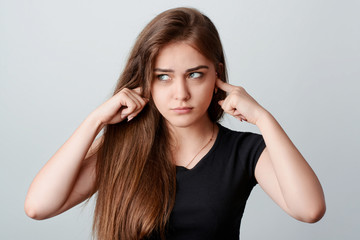 Young annoyed girl in black shirt sticking fingers in ears and looking right to the source of...