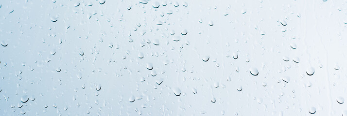 Wide picture of rain drops on a window glass surface against a white background of rainy clouds.