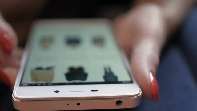 Woman looks at the goods  in the online clothing store. People using Smartphone, online shopping. Girl is choosing a shirt. Close up. 4K UHD. Screen is blurred.