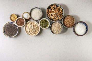 Variety of raw uncooked grains superfood cereal chia, linen, sesame, mung bean, walnuts, tapioca, wheat, buckwheat, oatmeal, coconut, rice in ceramic bowls grey spotted background. Flat lay space