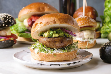 Variety of homemade classic, vegan and mini burgers in wheat and black buns with beef and veal cutlets, portobello mushroom, avocado on white ceramic board over white marble table. With glass of cola.