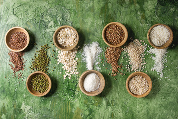 Fototapeta na wymiar Variety of raw uncooked grains superfood cereal linen seeds, sesame, mung bean, wheat, buckwheat, oatmeal, coconut, rice in wooden bowls over green texture background. Flat lay, space