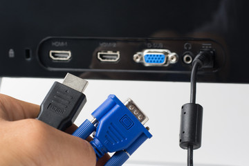 Man`s hand holds HDMI and VGA cables against a monitor with ports. Choise between modern HDMI and...