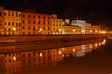 Illuminated buildings at night on the banks of the Arno river in Pisa and its reflection in the black waters.