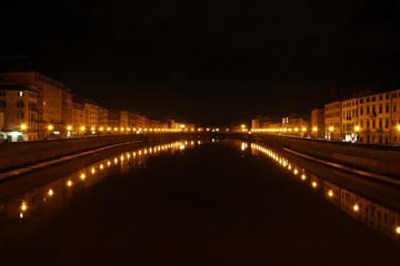 Naklejka na ściany i meble Illuminated buildings at night on the banks of the Arno river in Pisa and its reflection in the black waters.