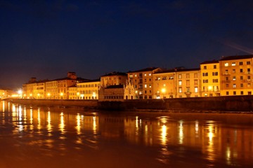 Illuminated buildings at night on the banks of the Arno river in Pisa and its reflection in the black waters.