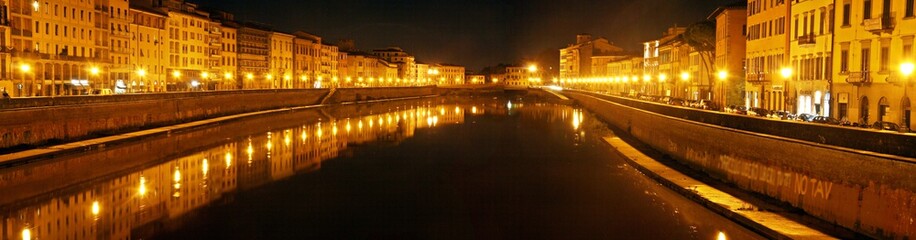 Fototapeta na wymiar Panoramic View of the Illuminated buildings at night on the banks of the Arno river in Pisa and its reflection in the black waters.
