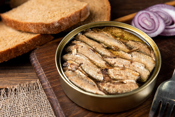 Sprats in tin can and bread on dark wooden background.