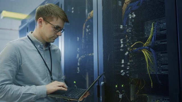 In Data Center IT Engineer Stands Before Working Server Rack Doing Routine Maintenance Check, Diagnostics Using Laptop. Visible Computer Hardware Equipment, Broadband Fiber Optic Cables and LED Lights