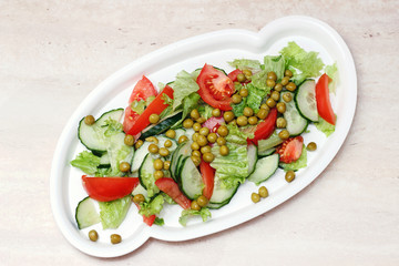 Original salad for healthy nutrition of cucumbers, tomatoes, lettuce and green peas on a white plate with olive oil, sprinkled with pepper on a marble table