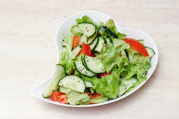 Original salad for healthy nutrition of cucumbers, tomatoes, lettuce on a white plate with olive oil, sprinkled with pepper on a marble table, a heart-shaped plate