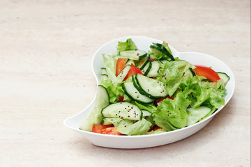Original salad for healthy nutrition of cucumbers, tomatoes, lettuce on a white plate with olive oil, sprinkled with pepper on a marble table, a heart-shaped plate