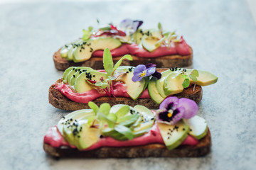 Low angled shot of avocado rye toast on beetroot hummus with edible purple pansy flowers, micro greens and nigella seeds or onion seeds, on marble background. soft focused background