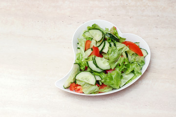 Original salad for healthy nutrition of cucumbers, tomatoes, lettuce on a white plate with olive oil, sprinkled with pepper on a marble table, a heart-shaped plate