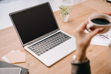 cropped view of businesswoman holding cup of coffee while sitting at workplace with laptop