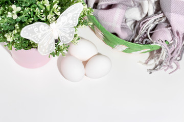 Easter white chicken eggs and green spring plant in flower pot, decorative butterfly and a basket with woolen scarf on white table with copy space