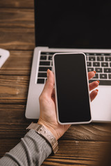 cropped view of businesswoman holding smartphone at workplace with laptop