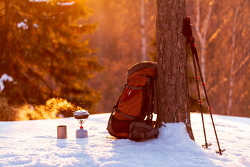 Fototapeta na wymiar kettle with steaming tea, thermo cup, backpack, trekking poles in the winter forest at sunset. The background is blurred. Sun glare.