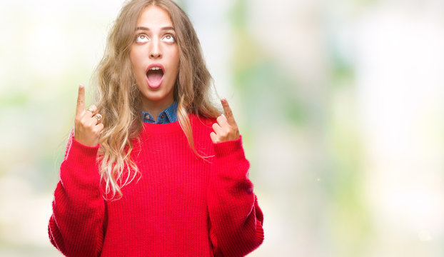 Beautiful young blonde woman wearing winter sweater over isolated background amazed and surprised looking up and pointing with fingers and raised arms.