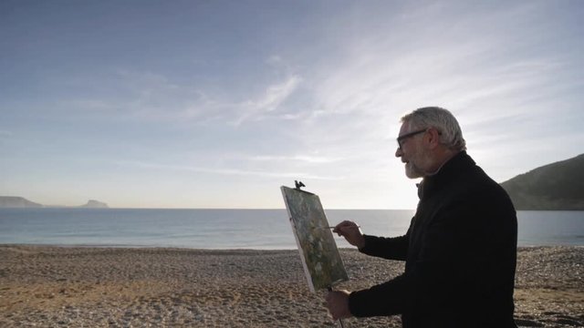Senior man paints a picture on the beach. Elderly male artist makes the finishing touches on the modern abstract painting against raising sun over seashore. Morning sun reflected in the calm sea water