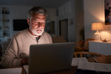 Senior Hispanic man checking his finances online at home using a laptop computer at night