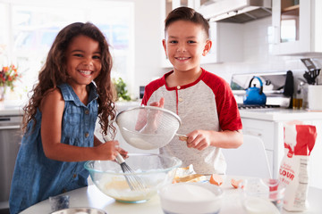 Brother and sister preparing cake mixture together at the kitchen table looking to camera, waist up