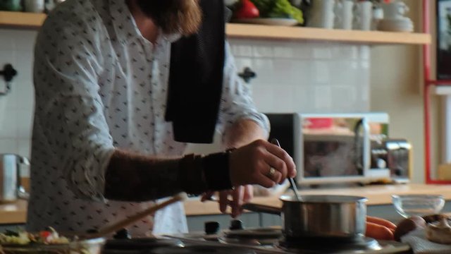 A cheerful man cooks alone in the kitchen, enjoying the process. Watching the dish on the stove and making a salad. Man is cooking. Charismatic male hipster is cooking in the kitchen.