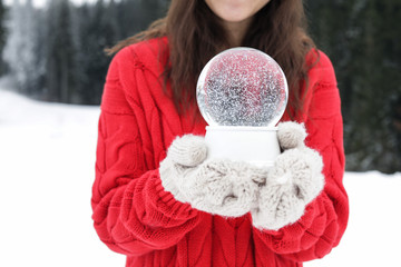Woman with knitted mittens holding snow globe outdoors, closeup. Winter vacation