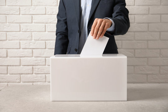 Man putting his vote into ballot box on table against brick wall, closeup