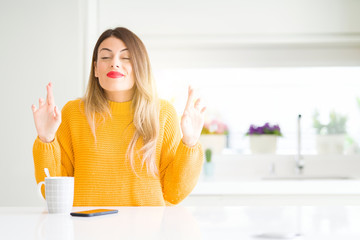 Young beautiful woman drinking a cup of coffee at home smiling crossing fingers with hope and eyes closed. Luck and superstitious concept.