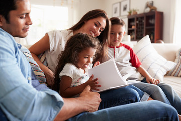 Young Hispanic family of four sitting on the sofa reading book together in their living room