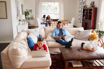 Pre-teen boy lying on sofa using laptop, dad using tablet, mum, sister and grandma in the background