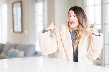 Young beautiful woman wearing winter fur coat at home looking confident with smile on face, pointing oneself with fingers proud and happy.