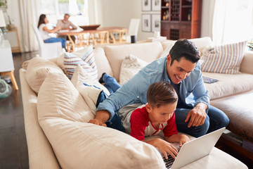 Pre-teen boy lying on sofa using laptop with his dad. Mum and grandad at the table in the background