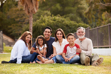 Three generation Hispanic family sitting on the grass in the park smiling to camera, selective focus