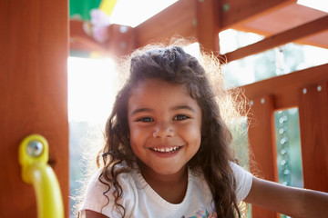 Young Hispanic girl playing on a climbing frame in a playground smiling to camera, backlit, close up - Powered by Adobe