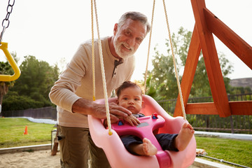 Senior Hispanic grandfather pushing his baby grandson on a swing at a playground in the park