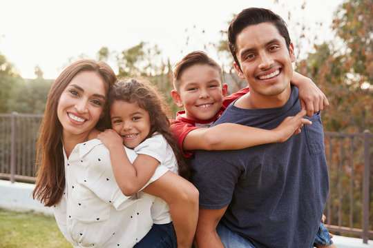 Young Hispanic parents piggyback their children in the park, smiling to camera, focus on foreground