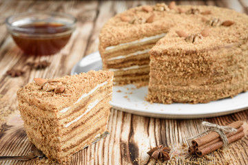 Homemade sweet layered honey cake with nuts on white plate on wooden table with bowl of honey, cinammon and badian.