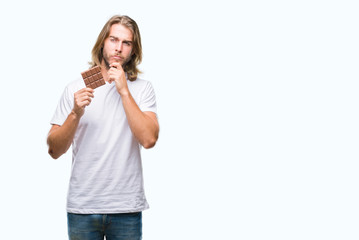 Young handsome man with long hair eating chocolate bar over isolated background serious face thinking about question, very confused idea